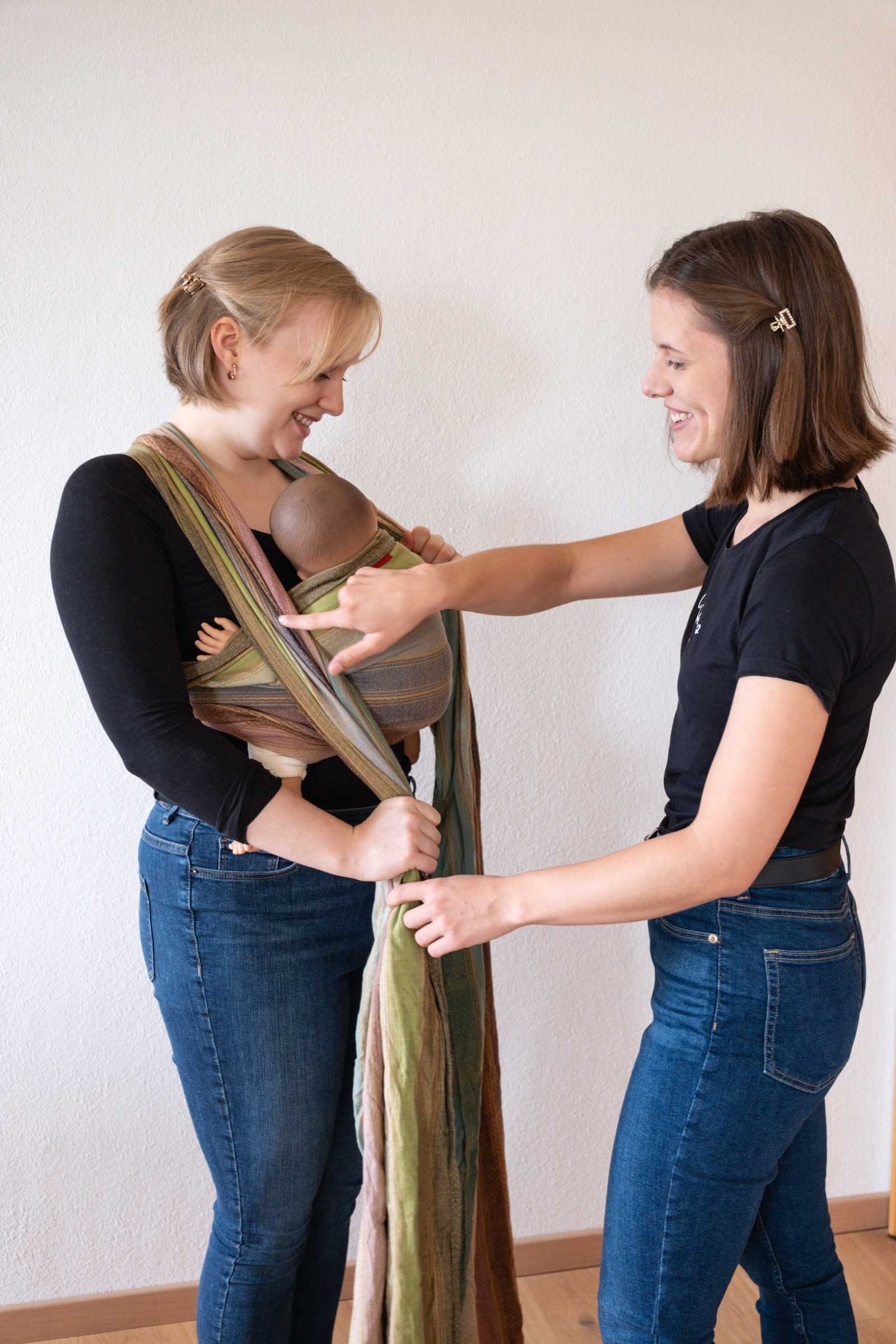 Sarah und Maya zeigen gemeinsam das Tragen mit einem Tragetuch und einer Babypuppe