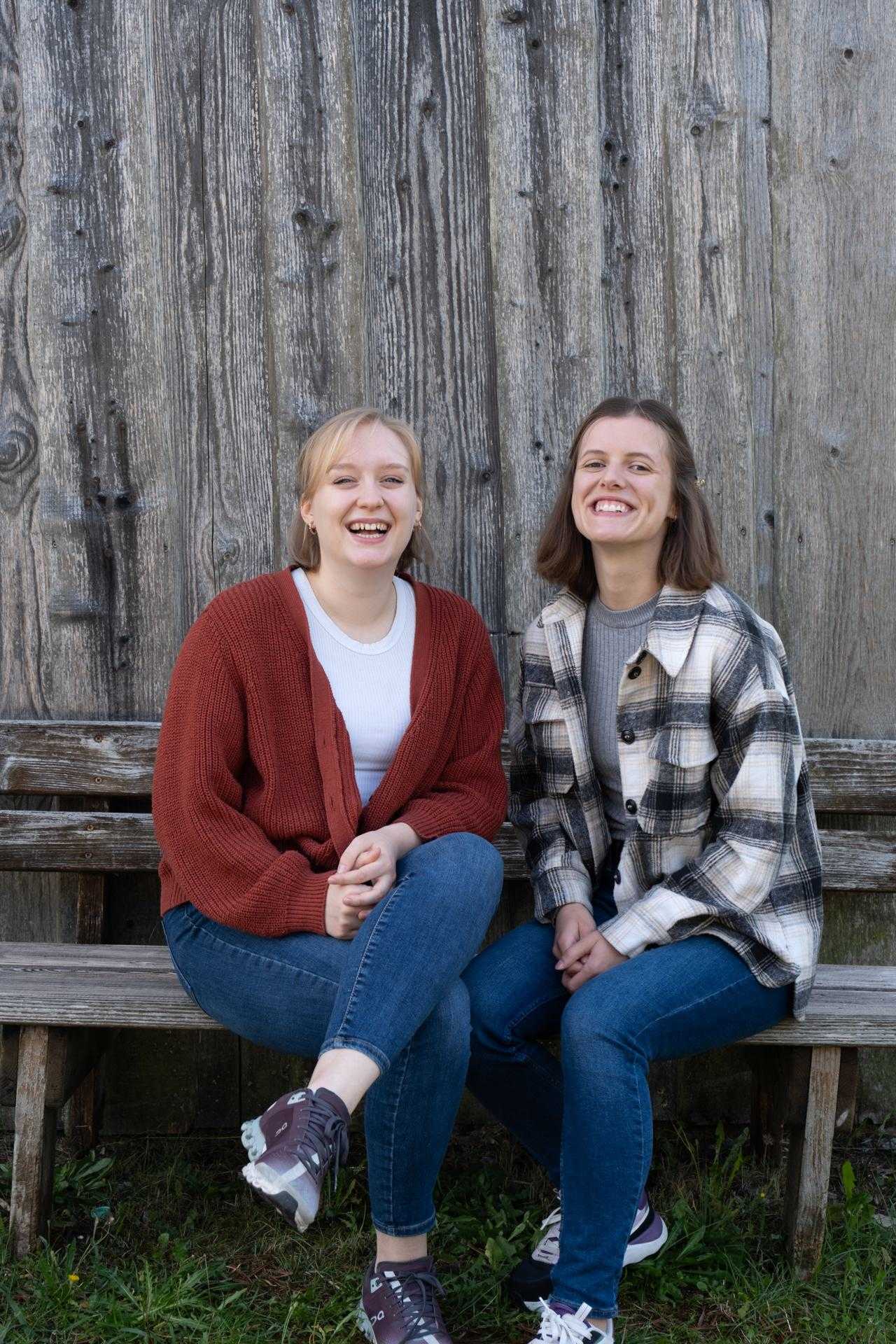 Maya und Sarah sitzen lachend vor einer Holzwand