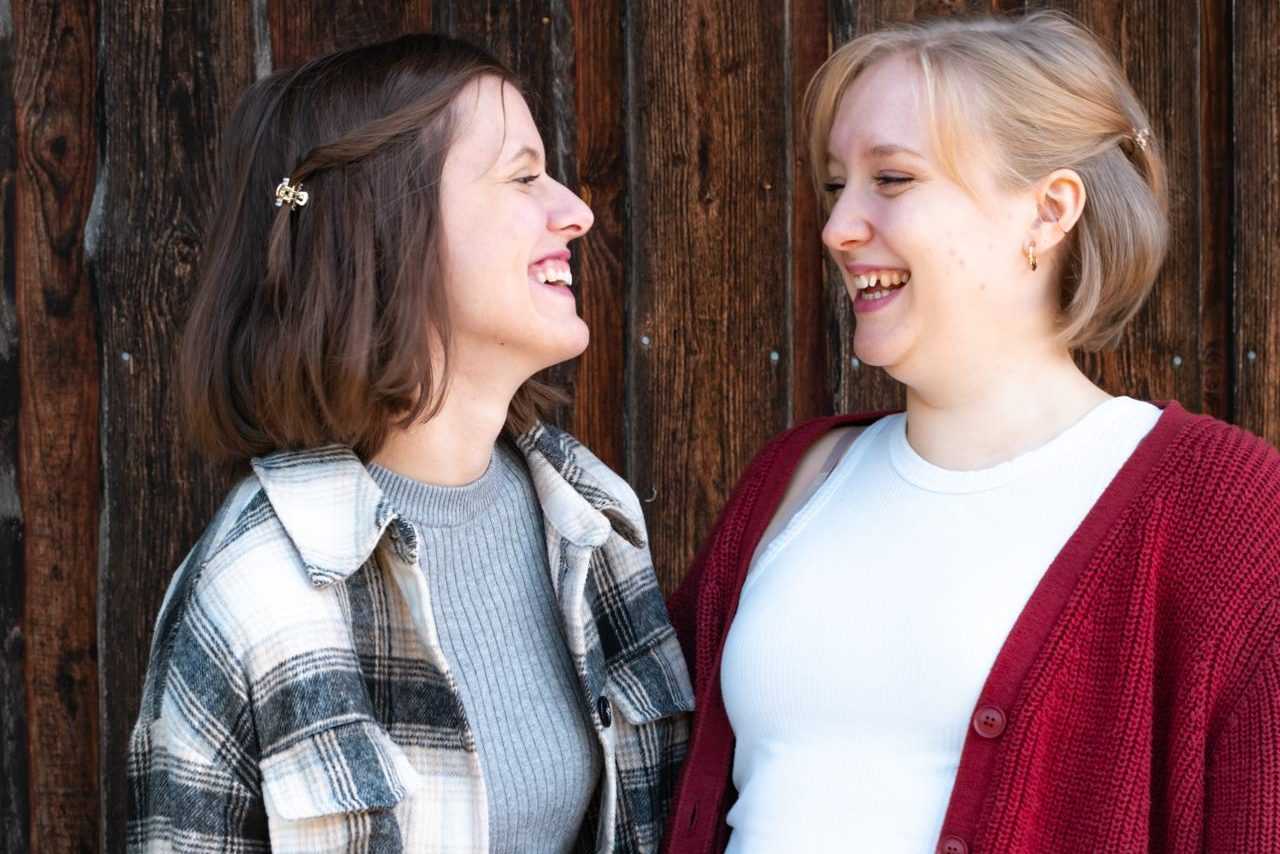 Maya und Sarah lachen sich vor einer Holzwand an